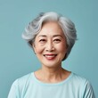 Portrait of a smiling senior Asian woman with short gray hair on a blue background. Happy old Chinese woman with a smile in a blue shirt looking at camera. Aged Japanese woman with shiny white teeth.