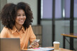 © Songsak C - Portrait of a beautiful confident businesswoman using a laptop computer holding a mobile phone sitting in a modern office. Smiling African American freelancer working online from home.