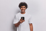 Fototapeta  - Indoor shot of positive handsome Hindu man using smartphone texting to family sending sms browsing web checking social network on mobile device dressed in casual t shirt isolated on white background