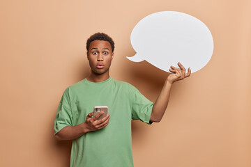 Wall Mural - Studio photo of young confused African guy holding white speech bubble with blank space for advertisement shocked reading news on his smartphone isolated on beige background. People emotions concept