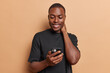 © WHstudio Leushin N - Pleased dark skinned man keeps hand on neck smiles pleasantly holds mobile phone watching video using entertaining mobile application dressed in casual black t shirt isolated over brown background.