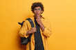 © WHstudio Leushin N - Shocked curly haired male model with backpack on shoulder stares impressed wears shirt keeps hand on chin cannot believe in shocking news isolated over vivid yellow background. Lifestyle concept