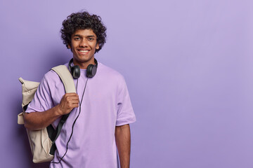 Wall Mural - Professsional studio close up of young smiling happy hindu man isolated on purple background wearing casual purple tshirt holding big white leather bag on his left shoulder with headphones on neck