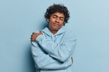 Waist up shot of pleased Hindu man with curly hair embraces himself keeps eyes closed dreams about something dressed in comfortable sweatshirt isolated over blue background. Love yourself concept