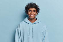 Portrait Of Handsome Curly Haired Young Hindu Man Smiles Pleasantly Dressed In Casual Hoodie Expresses Positive Emotions Poses Against Blue Studio Background. Monochrome Shot. Youth Concept.
