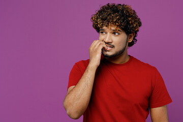 Wall Mural - Young minded pensive sad confused thoughtful Indian man he in red t-shirt casual clothes look aside on area biting nails fingers isolated on plain purple background studio portrait Lifestyle concept