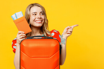 Traveler woman wears red dress casual clothes hold bag suitcase passport ticket point aside isolated on plain yellow background Tourist travel abroad in free time rest getaway Air flight trip concept