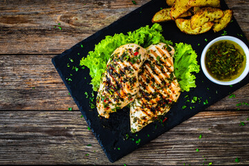 Wall Mural - Grilled chicken breasts with Argentinian chimichurri sauce,  baked potatoes and fresh vegetables on wooden table
