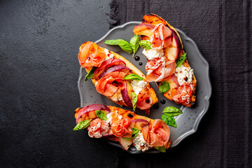 Poster - A dark grey plate with with open sandwiches, snack. Toastet bread, griddled peach, parma ham or prosciuto and robiola cheese, with balsamic vinegar and basil. Black background, top view.