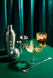 Splash in champagne glass, cocktail glass, shaker, cocktail mixing spoon and jigger on dark green table in disco club atmosphere