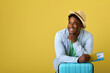 © Aleksandr - Dreaming African-American man in hat with plane ticket in his hand leaned on blue suitcase. A cheerful dark-skinned guy on a yellow background with a ticket in his hand dreams of an upcoming trip