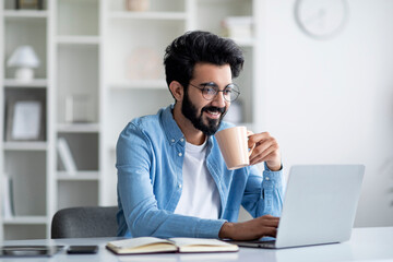 Wall Mural - Young Smiling Indian Freelancer Guy Using Laptop And Drinking Coffee At Home