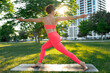 Young woman wearing in pink sportwear doing yoga on a black mat in morning park. Warrior Pose