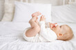 © Any Grant - smiling baby 6 months old blond boy lies on a large bed in a bright bedroom and plays with his legs in a cotton bodysuit, the concept of children's goods