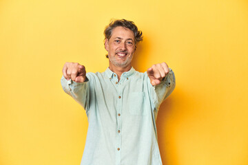 Wall Mural - Middle-aged man posing on a yellow backdrop cheerful smiles pointing to front.