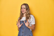 © Asier - Young blonde Caucasian woman in denim overalls posing on a yellow background, rejecting someone showing a gesture of disgust.