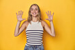 © Asier - Young blonde Caucasian woman in a white tank top on a yellow studio background, showing number ten with hands.