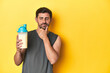Fit man with protein shake, yellow studio background looking sideways with doubtful and skeptical expression.