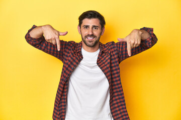 Caucasian man in red checkered shirt, yellow backdrop points down with fingers, positive feeling.