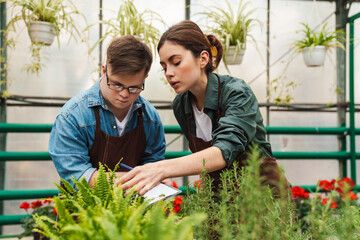 Wall Mural - Man writing down notes in clipboard while woman gardener teaching him to handle with flowers in greenhouse