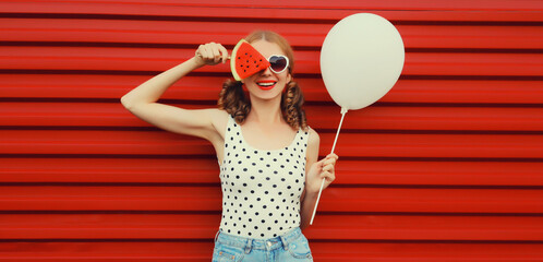 Happy cheerful young woman with balloon and fresh fruit lollipop or ice cream shaped slice of watermelon wearing white heart shaped sunglasses on red background