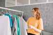 © Shi  - Clothing, purchasing, style concept. Happy blonde haired woman chooses clothes at home wardrobe, holds comfortable jumper on hangers, outfits on racks in background, messages via mobile phone. High