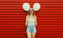 Happy Cheerful Young Woman Having Fun With Balloon Wearing White Heart Shaped Sunglasses, Shorts On Red Background