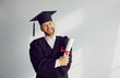 Portrait of happy male graduate student standing with diploma. Positive young man wearing graduation robe and mortarboard smiling at camera standing at gray wall. Graduation, education concept