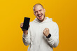 Excited young man screaming and showing empty screen smartphone while celebrating victory with raised fist, isolated over yellow background