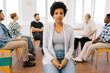 © dikushin - Portrait of pretty African young woman looking at camera with serious expression during group therapy session, multicultural and different ages people sitting in circle on background.