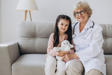 Portrait Of Senior Nurse Wearing White Coat, Embracing Shoulders Of Happy Little Preschool Patient. Smiling Small Girl Holding Favorite Toy, Posing For Photo With Attractive General Practitioner.