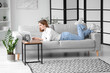 Pretty young woman lying on grey sofa and using modern laptop in light living room