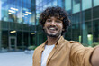 © Tetiana - A young, successful and smiling Indian male businessman is standing outside an office center and talking on an online call, smiling at the camera, recording a video