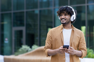 Happy young Indian man listening to music, podcast on his phone. Standing outside wearing headphones and looking away with a smile