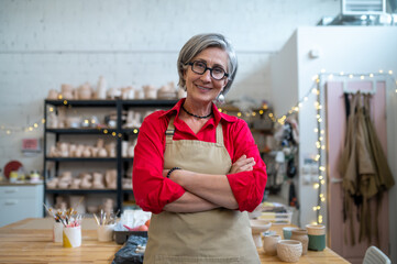 Wall Mural - Happy senior woman successful self-employed potter wearing apron behind table with pottery tools.