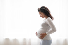 Beautiful Pregnant Black Woman Hugging Tummy While Standing Near Window At Home