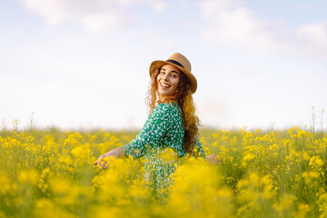 Beautiful woman in bright dress and elegant hat walks and has fun in rapeseed field. Smiling female tourist walking through flowering field, touching yellow flowers. Nature, rest. Summer landscape.