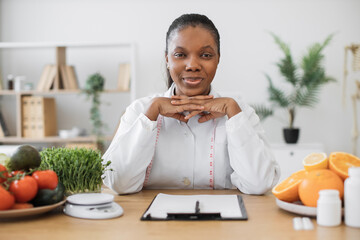 Serious multiethnic woman in doctor's coat carrying out professional activity as nutrition specialist in clinic. Confident health professional thinking over meal plans of clients at office desk.