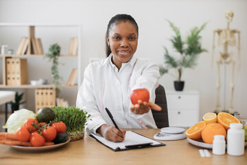 Wall Mural - Happy multicultural female in white coat reaching out hand with fresh apple while working in dietitian's office. Efficient health professional encouraging to overall health and well-being via eating.