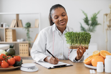 Wall Mural - Positive female nutritionist with young sprouts in hand creating meal diet at writing desk in doctor's office. African american specialist with measuring tape recommending clients slimming foods.