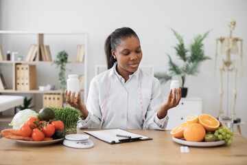 Canvas Print - Focused female nutritionist holding two medicine bottles while working at office desk with products on it. Multiethnic health professional choosing food supplement for balancing diet of clients.