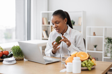 Sticker - Charming adult lady in lab coat showing avocado to front camera of computer in professional office of clinic. Multicultural specialist in nutrition counseling patient online about benefits of fruit.