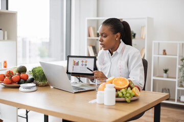 Canvas Print - Confident lady in doctor's coat getting presentation on laptop via conference call during working hours in clinic. Efficient multiracial specialist in food providing nutritional guideline via graphs.