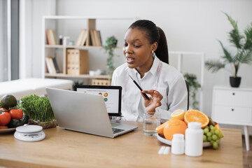 Focused multicultural expert with meal plan on tablet conducting web meeting using portable laptop at work. Skilful female nutritionist explaining details of slimming program for client online.