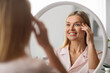 © Prostock-studio - Cheerful mature woman looking at mirror at home, applying eye cream