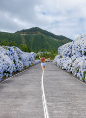 Wall Mural - Azores, Young woman jumping happy in the middle of road with blue hydrangea at the roadside in Lagoa Sete Cidades 