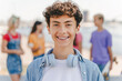Authentic portrait of smiling teenage boy with braces wearing headphones looking at camera standing on the street with friends on background. Positive lifestyle, summer concept