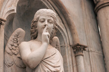 Religious Statue Figure Of An Angel Making Silence On A Grave In An Outdoor Cemetery