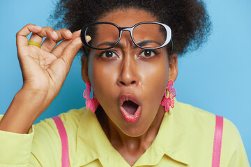 Close-up face of surprised black girl, looks into camera with opened mouth, lifting her glasses to forehead with one hand, interesting fact concept, copy space