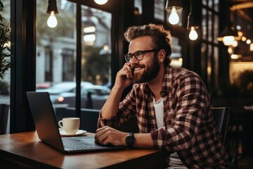 Portrait of a handsome bearded man in glasses and a plaid shirt sitting in a cafe with a laptop and talking on the phone. Generative AI